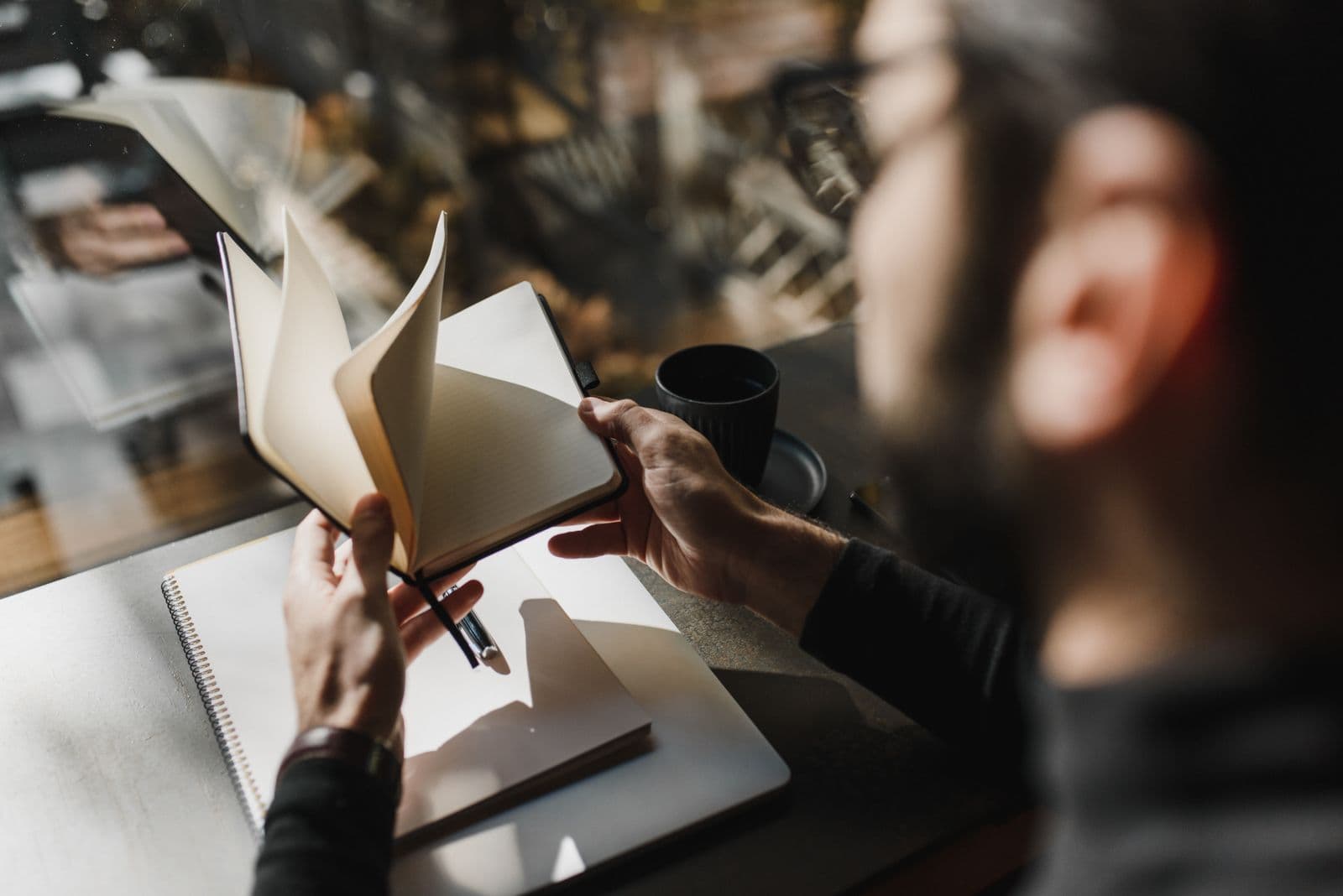 Someone paging through a notebook by a bright window, coffee in hand
