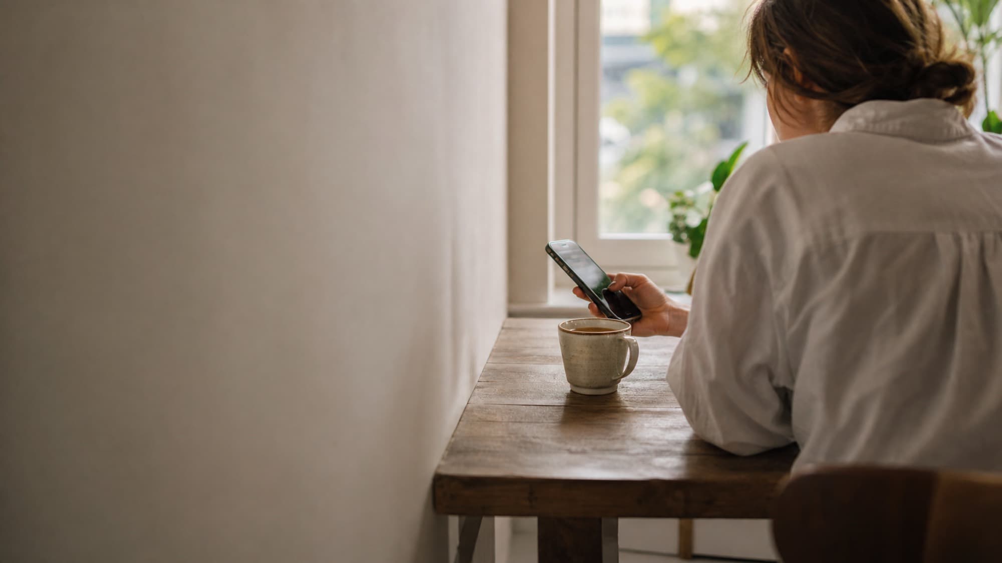 A person at a sunny window glancing at their phone, a coffee on the table